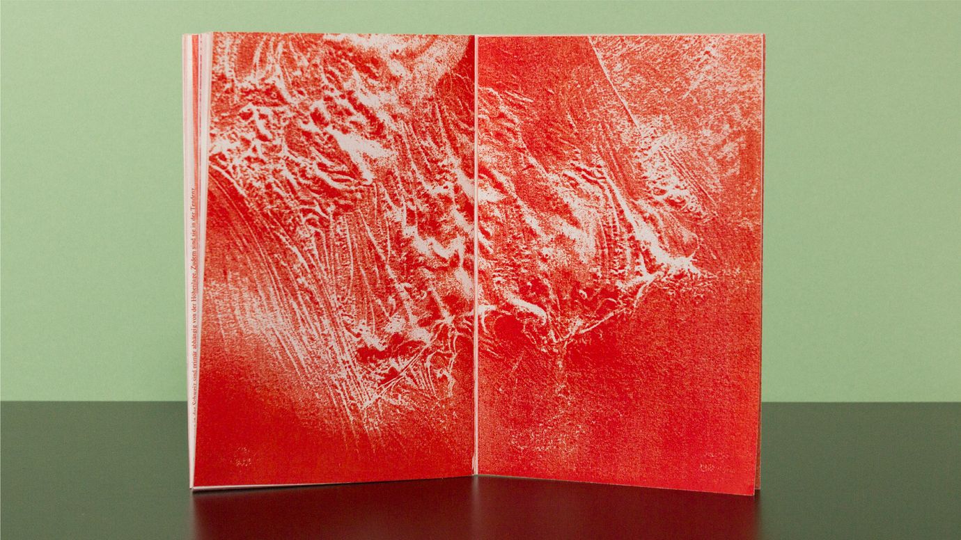 Two-page spread from a copy magazine. Full-bleed, semi-abstract image of a mountain topography, printed in red. The magazine is standing on a dark table. Neutral light green background.