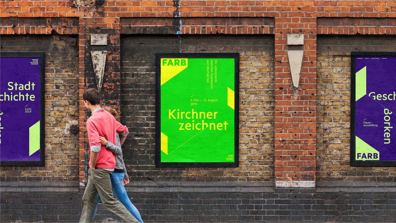 Posters on a brick wall. Passers-by. The posters are brightly colored and feature geometric elements. The letters FARB are placed in one corner on a flat element that suggests a three-dimensional shape protruding into the poster. The central poster is titled “Kirchner Zeichnet” (Kirchner draws).